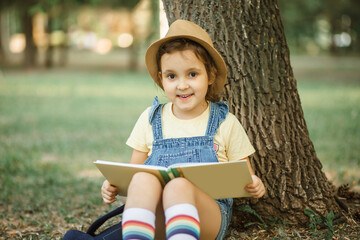 Little cute girl kid sitting under the tree on summer in the park outside and reading a book.