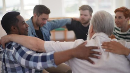 Back view of mature male therapist and group of multi-age patients putting hands on each others shoulders sitting in circle during group therapy session as symbol of support. Shooting in slow motion.