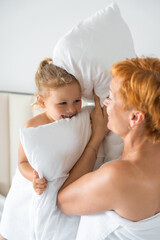 Young mother and little daughter having fun with pillows on bed