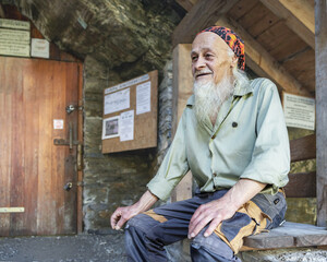 An old cave keeper with a gray beard and a smile sits at the entrance with a cigarette in his hand
