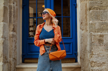 Fashionable happy smiling woman wearing trendy orange linen shirt, bandana, crochet top, denim skirt, blue sunglasses, holding stylish baguette bag, posing in street. Copy, empty space for text