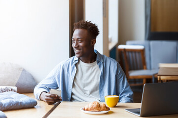 Fototapeta na wymiar Millennial African guy with smartphone and laptop sitting in cafe