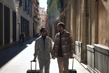 Real marriage of gay couple, holding hands, with a suitcase in the other, looking at each other with complicity, walking down the street. Concept lgtb, lgtbiq+, couples, in love, travel.