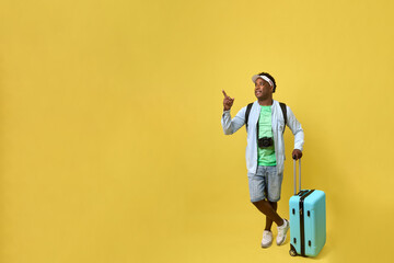 Fototapeta na wymiar Happy young man standing on a yellow background with a suitcase pointing to an empty space for a travel agency advertisement. A cheerful traveler with luggage is going on summer vacation