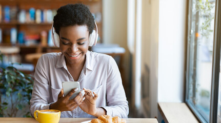Cheerful lady in headphones listening music on smartphone at cafe