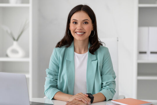 Positive confident young european lady manager in suit sit at table workplace in modern office