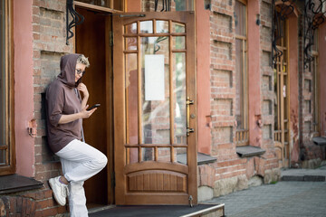 Caucasian woman in a casual wear stands propping up the wall outside and using her mobile, selected focus.