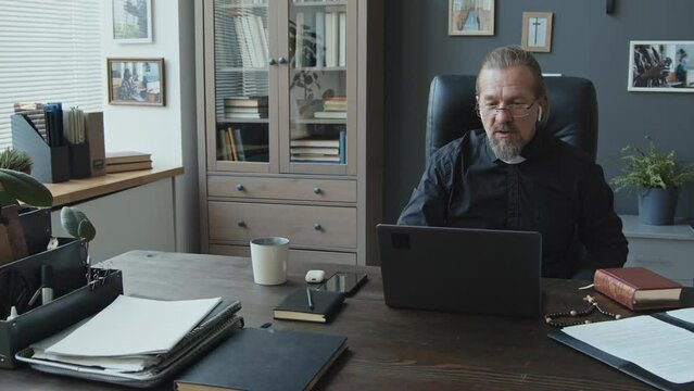 Modern mature Caucasian Catholic priest sitting at table in office discussing something with colleagues during video conference