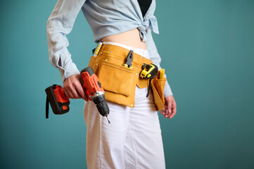 Fototapeta na wymiar Young female worker with tool belt holding an electric screwdriver in hands isolated on background. Pretty caucasian female with tools planning new project. Portrait in studio. Crop view.