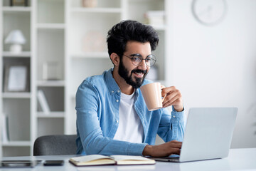 Young Smiling Indian Freelancer Guy Using Laptop And Drinking Coffee At Home