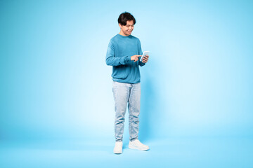 Fototapeta na wymiar Korean guy with cellphone standing texting over blue studio background