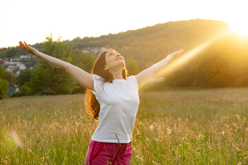 A woman stands in a field in nature with her hands raised in the rays of the setting sun in summer.