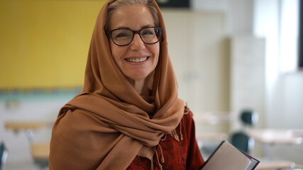 Portrait of attractive muslim teacher woman with headscarf in an empty classroom holding books.