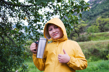 A cute young little boy in yellow jacket is drinking tea and showing a thumb on mountains background.