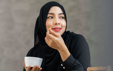 Beautiful Muslim woman wearing traditional clothes or costume, holding small cup, eating snacks at home, smiling with happiness. Lifestyle and Religious Concept.