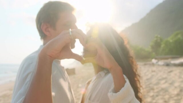Young enamored couple Indian woman and Caucasian man make heart shape from fingers together showing love and rejoicing at beginning of romantic relationship or first date stands on sea beach.