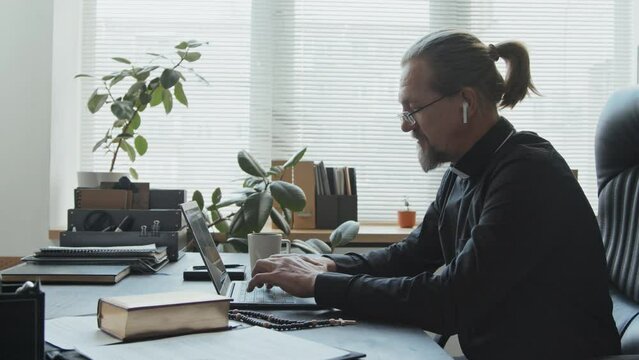 Side view shot of modern Catholic priest wearing earbuds sitting in office texting something on laptop and drinking hot tea