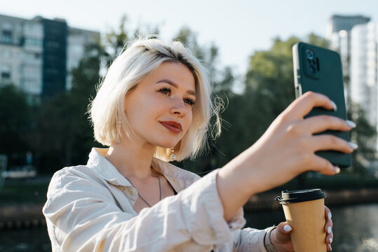 Young woman taking selfie on smartphone, cute blonde teenager with cup of coffee using phone camera outdoors
