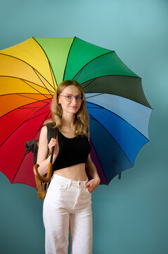 Happy caucasian female in black top and white pants under rainbow colored umbrella posing at studio background, free copy space. Photo portrait of cute girl under umbrella.