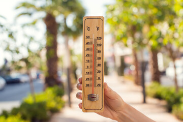 Hot weather. Thermometer in hand in front of street with trees and palms during heatwave. High...