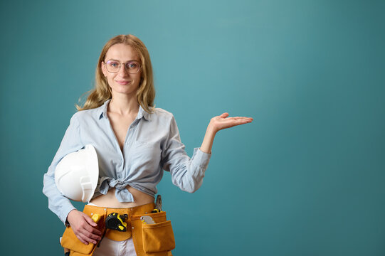 Portrait Of Young Female Worker With Tool Belt Showing A Product With Her Hand Isolated On Background. Pretty Caucasian Female With Tools Planning New Project. Portrait In Studio. Copy Space.