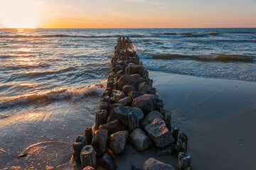 Perspective view of an old sea breakwater made of logs and stones. The waves coming from the sea are illuminated by the setting sun. Seascape. - Powered by Adobe