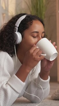 Vertical video. Cozy relax. Morning coffee. Hygge lifestyle. Dreamy woman drinking tea listening music in headset resting on floor home room.
