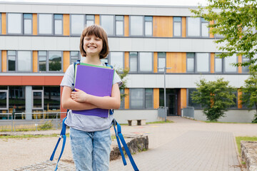 Portrait of a happy teenage girl standing with a backpack on her back in front of the school with textbooks in her hands. Back to school, start of the school year.