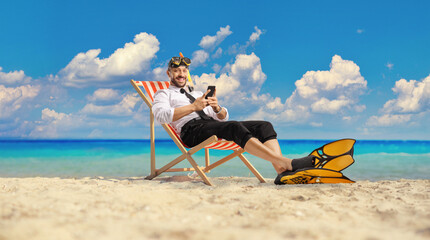 Businessman with snorkelling fins and mask sitting on a bech chair by the sea and using a mobile phone
