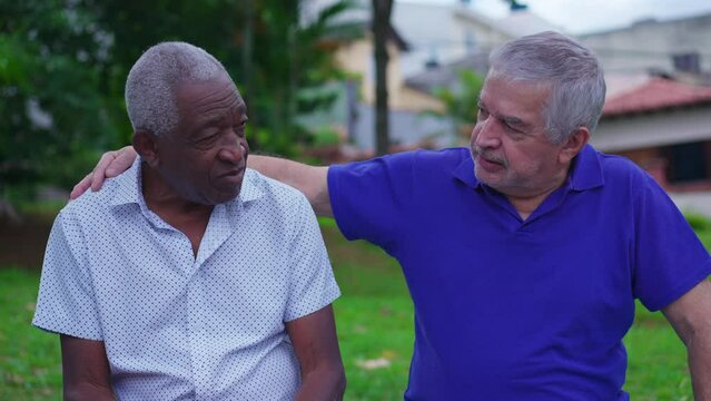 Two diverse senior friends sharing moment of camaraderie sitting on park bench, older caucasian friend putting hand over African American colleague shoulder in conversation