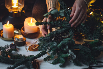 Young woman in knitted sweater doing handmade Christmas wreath from natural materials: fir tree branches, dry citrus slices, cones, cinnamon sticks. Wooden table, cozy light, home atmosphere