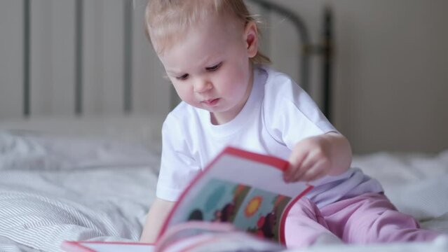  little baby girl reading, looking at pictures in book on bed at home, An inquisitive baby wants to read new fairy tale