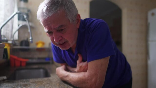 Senior man suffering heart attack alone at home leaning on kitchen sink and falling to the floor in dramatic acting. Elderly person struggling with illness