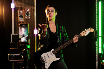 Obraz premium Rockstar blowing air kisses in front of smartphone camera while playing at electric guitar in sound studio, working at rock album using electricinstrument. Rebel musician recording heavy metal song