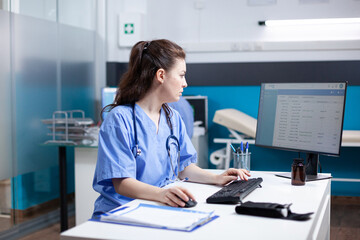 Caucasian nurse working at desk, inserting data in appointment table list. Concentrated adult woman worker wearing scrubs and stethoscope in clean, modern medical office, operating computer