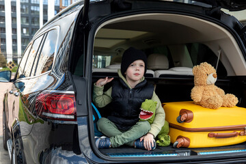 Adorable little kid boy sitting in car trunk just before leaving for vacation with his parents. Happy child with suitcases and toys going on journey