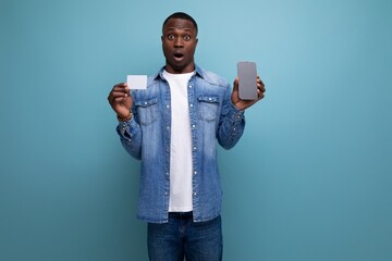 handsome surprised young attractive african man dressed casually in denim clothes holding credit card and smart phone on background with copy space
