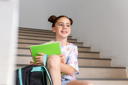 Portrait of a smiling red haired girl with a backpack and holding notebooks sitting on the school stairs, back to school concept.