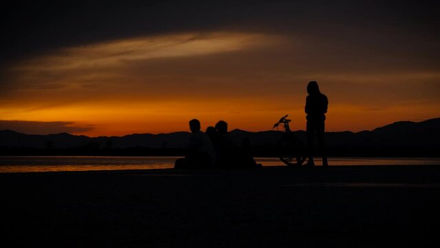 Silhouette of teen group during sunset. Silhouettes of boys and girls relaxing with phone on the beach by waterfront during vacation time.