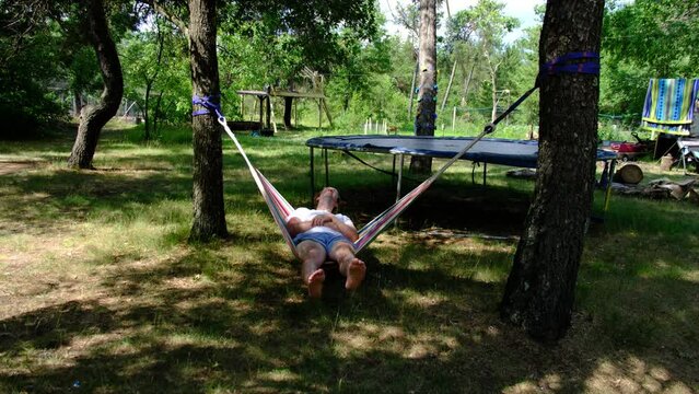 Man in white shirt and blue shorts being barefoot swing in small hammock in between two trees ij front of trampoline.