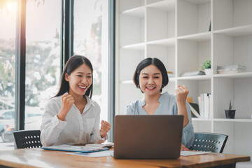 Two businesswomen show joyful expression of success at work smiling happily with a laptop computer in a modern office.