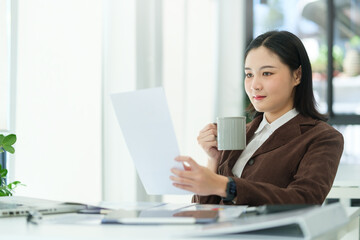Attractive businesswoman drinking coffee and reading marketing data paper report at desk.