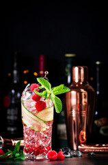 Rose garden alcoholic cocktail drink with dry gin, tonic, rose syrup, lychee juice, lemon, raspberry, cucumber and blackberry with ice. Black bar counter background, bar tools and bottles