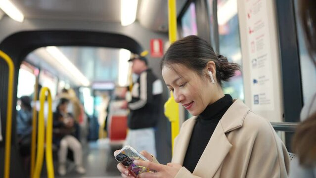 Asian woman using mobile phone and listening to the music on earphones during travel on train in the city. Attractive girl enjoy urban lifestyle in the city with using wireless technology on device.