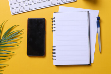 Blank page of a notebook for your text with black smartphone, laptop and palm leaf on yellow office...