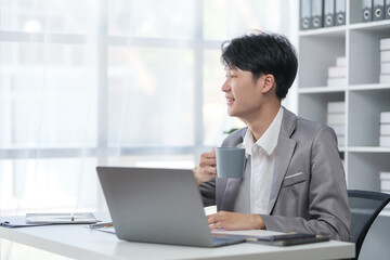 Fototapeta premium Relaxed asian man holding coffee cup and looking away while sitting in modern office.