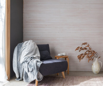 Grey armchair with throw rug in rustic room with small table and dried leaves in ceramic jug against white painted wood panelled wall (selective focus)