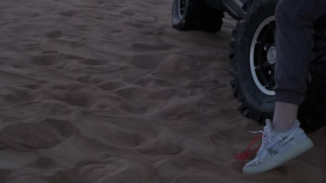 Young woman in sportswear standing next to buggy on desert sand dunes against sunset background