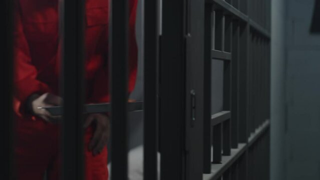 Elderly prisoner in orange uniform sits in prison cell. Prison officer gives tray of food through metal bars. Inmate eats dinner, serves imprisonment term for crime in jail or correctional facility.