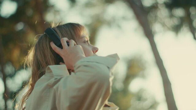 Girl puts headphones on head at golden hour, listens to podcast. Close side view